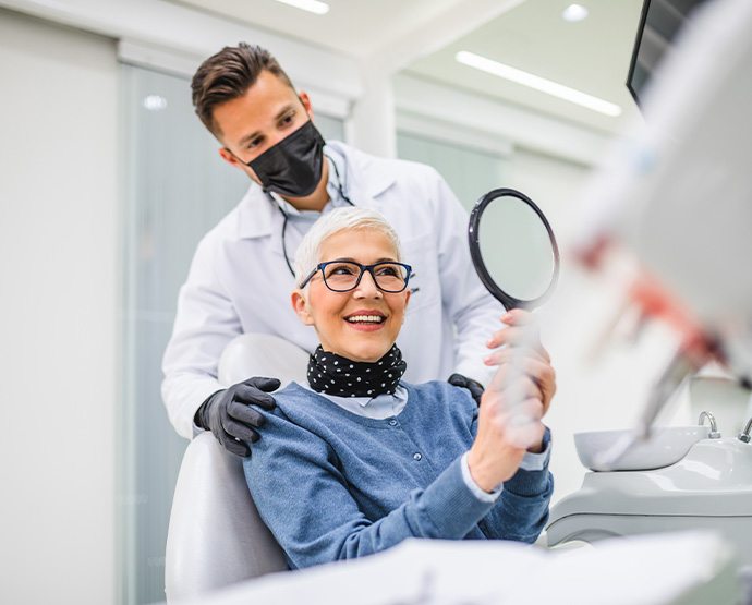 Woman smiling at reflection in handheld mirror with dentist